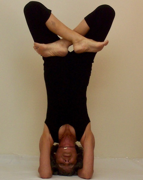 A thin woman in a black unitard does a headstand with legs crossed in a lotus position.