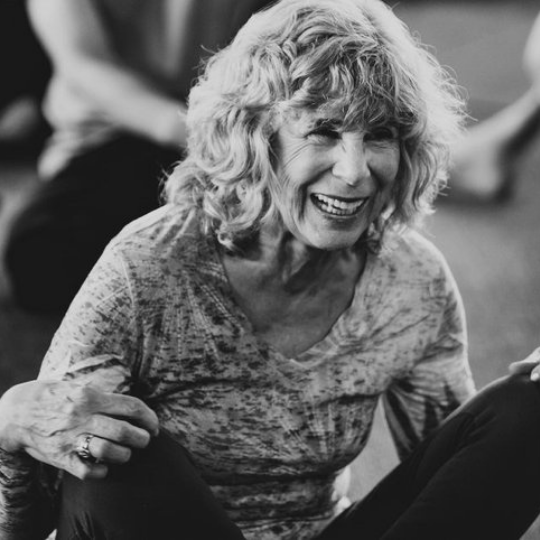 A thin woman with white hair kneels on a yoga mat gesturing to the side as she speaks to her yoga students.