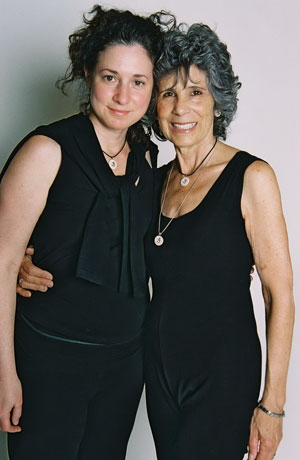A younger and an older Yoga teacher stand arm in arm, heads touching.