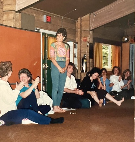 A line of seven seated women smiling, laughing, clapping hands. One stands in the centre smiling and looking on. 