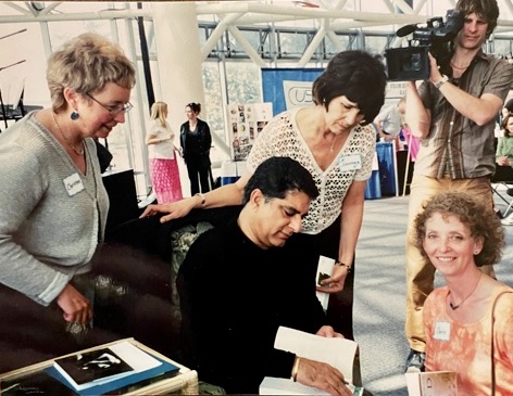 Three women look on as Deepak Chopra autographs a book. 