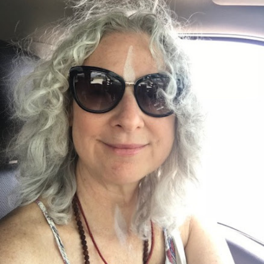 A white woman with curly grey hair, sunglasses, and sundress smiles from the seat of a car