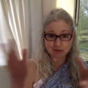 A lively woman with long grey hair and dark framed glasses tells a story, sitting in a trailer in front of a window. 