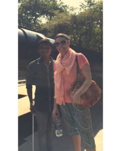 A white woman with short hair and pretty blouse and loose pants stands next to a South Asian man in button down shirt and cotton trousers. They stand in hot sun, in front of lush trees