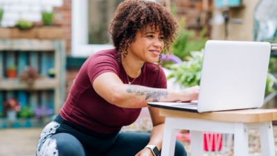 A yoga student with brown skin and medium length curly hair smiles as she taps on a laptop keyboard