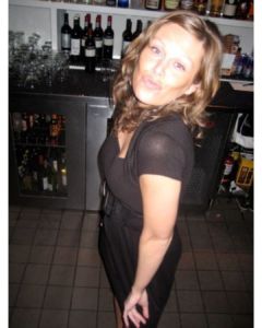 A beautiful woman poses for the camera in black dress, standing in front of a wall of wine and liquor bottles