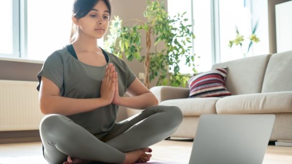 A yoga student of South Indian heritage faces a laptop, seated cross legged with hands in anjali mudra