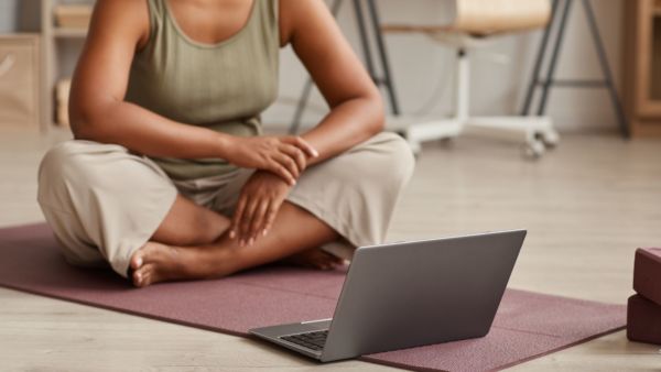 A yoga student with brown skin faces a laptop. Her face isn't visible.