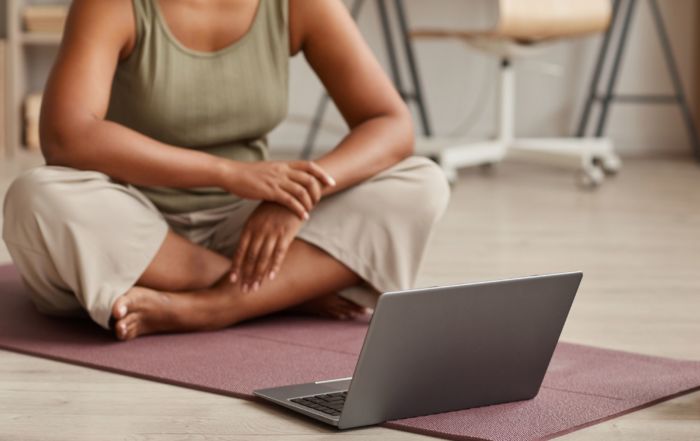A yoga student with brown skin faces a laptop. Her face isn't visible.