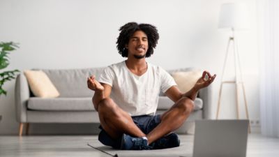 Black male yoga student sits cross-legged with hands in mudra on his knees smiling facing a laptop