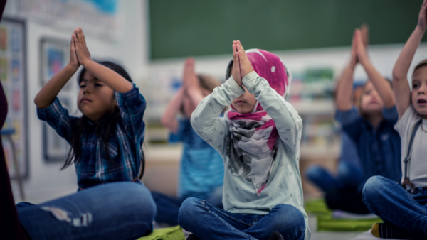 Small children sit in a classroom pressing palms together over their heads. One girl in the front wears a hijab. There is a girl with brown skin and long dark hair beside her. Behind her we see a girl with light skin and blonde hair.