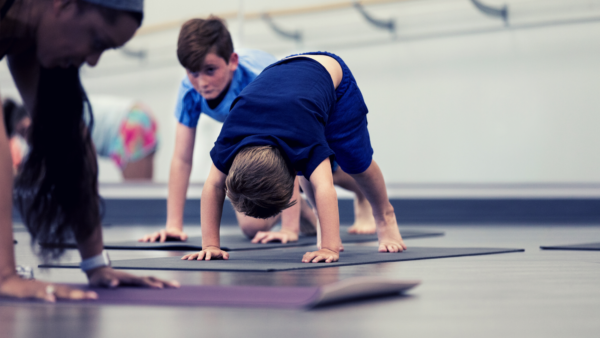 Two small boys try to do Downward Dog on yoga mats, a teacher is in the foreground