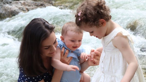 A white woman with long brown hair holds a baby. A small girl in a white dress and pink hair bows stands next to her. They're all focused on something at their feet and smiling. In the background, a rushing river. 